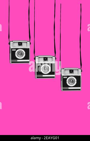 three gray and black retro film cameras, hanging from their strips, on a pink background, with some blank space on the bottom Stock Photo
