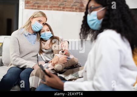 Beautiul skilled black-skinned woman in medical gown visiting sick teen girl which is ill at home and is under supervision of caring loving mother Stock Photo