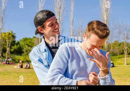 Casual Gay Couple Hugging in a Park and Scrtching Nose with People Picnic Background Stock Photo