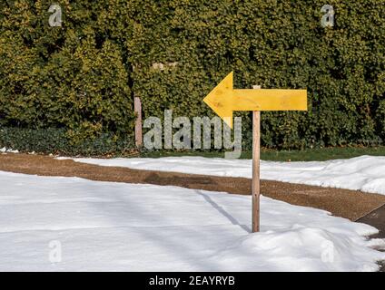 Yellow wooden arrow sign on post pointing left with green hedge background. Stock Photo