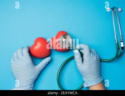 two hands hold medical stethoscope and red rubber heart on a blue background, close up Stock Photo