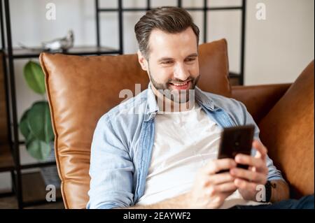 Cheerful young guy spends leisure scrolling news feed on the smartphone, a bearded guy holds mobile phone, looks at the screen, websurfing, shopping Stock Photo