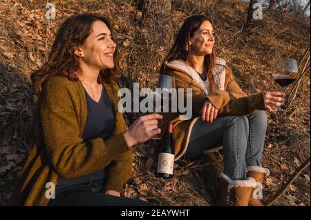 young women sitting in a forest looking at the sunset while drinking red wine. Friendship, togetherness concept. Stock Photo