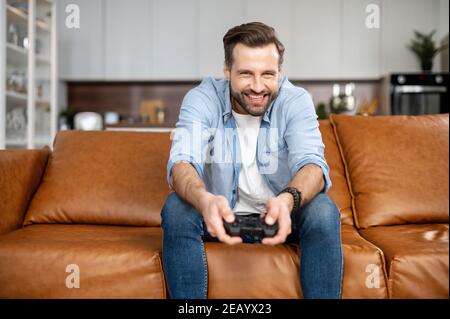 Handsome excited young guy spends leisure time playing video game, gambling guy with a joystick sitting on the couch, play on console at home Stock Photo