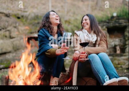 young happy women laughing, holding glass of red wine. Females warming next to the fire. Campfire, outdoors activities concept. Stock Photo