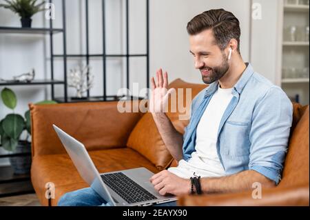 Smiling and cheerful young bearded man using a laptop for video call, video conference sitting at the comfortable couch at home. Handsome guy in Stock Photo