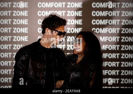 Indian Caucasian couple in stylish clothes with sunglasses smiling and looking each other. People standing on black background and illuminated by ligh Stock Photo