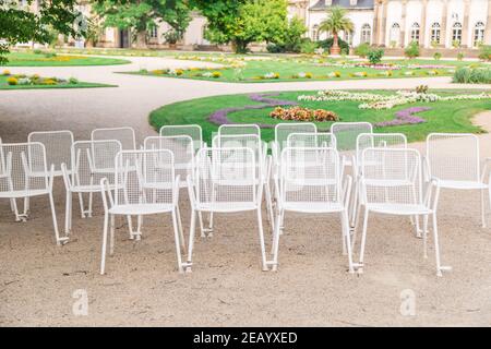 Rows of white metal chairs in summer park with paths and flower beds Stock Photo