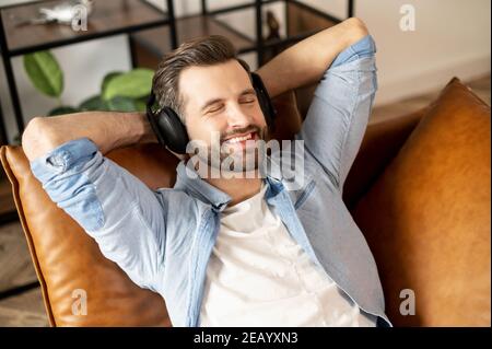 Handsome smiling guy wearing headphones enjoys music, attractive bearded man rests with a favorite tracks, lying on the comfortable sofa with eyes Stock Photo