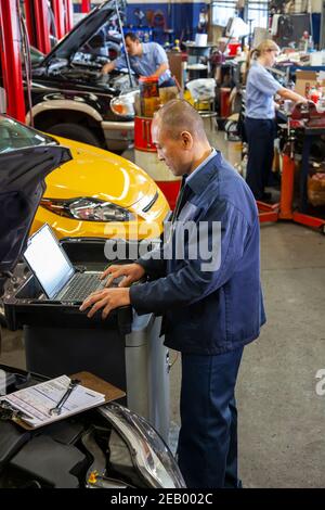 Pacific Islander mechanic typing on a laptop while co-workers work on cars in background Stock Photo