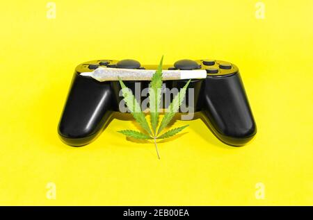 Video games and cannabis: game controller with marijuana leaf and big cannabis joint isolate on yellow background. Stock Photo