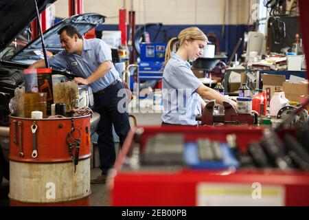 Caucasian female mechanic works on engine in auto repair shop with Hispanic coworker in background Stock Photo