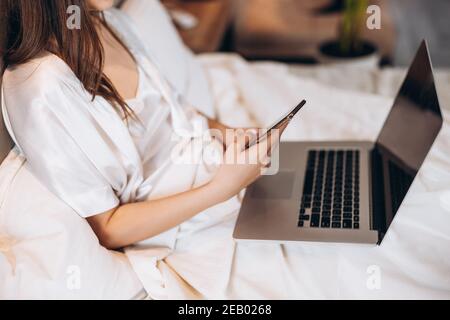 laptop and phone in the hands of a woman without a face. laptop and phone on the background of white pajamas. work at home Stock Photo