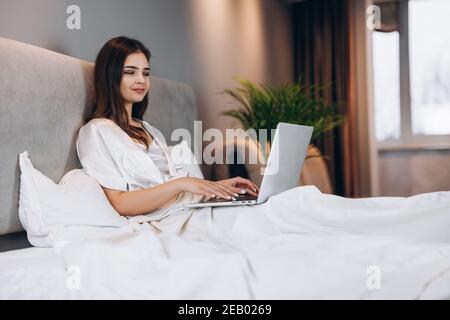 Young beautiful woman with laptop in bed. Brunette model with laptop in silk white pajamas. A woman works at a computer in her bedroom at home Stock Photo