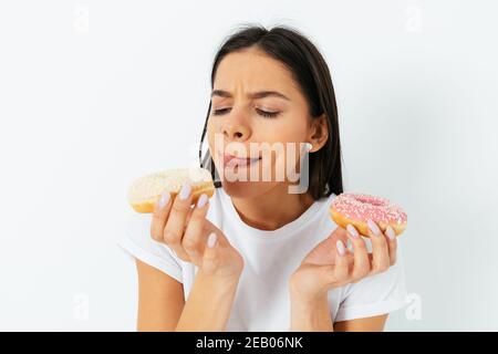 Young beautiful woman wants to eat donuts, looks at them licking her lips, isolated on white studio background. Stock Photo