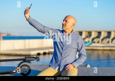 Mature man takes selfie with mobile phone. He is sitting and has an electric scooter. In the background you can see the sea. There is sunset light. Ho Stock Photo