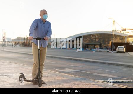 mature man wearing a mask uses an electric scooter in the city. He wears blue shirt and ochre pants. There is sunset light. Horizontal photo Stock Photo