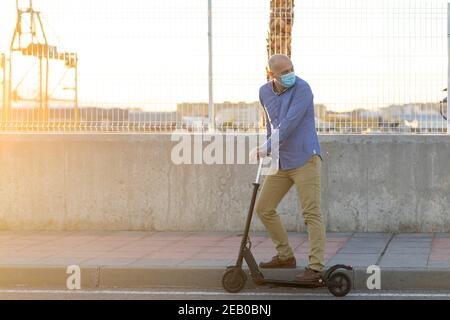 mature man wearing a mask uses an electric scooter in the city. He wears blue shirt and ochre pants. There is sunset light. Horizontal photo. looking Stock Photo