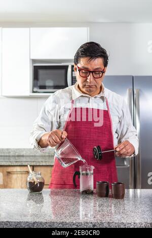 Vertical shot of man in red apron and mask making fresh coffee in a French press. Stock Photo