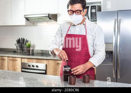 Horizontal shot of man in red apron and mask making fresh coffee in a French press next to two espresso cups in a kitchen. Stock Photo