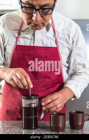 Vertical shot of man in red apron making fresh coffee in a French press next to two espresso cups. Stock Photo