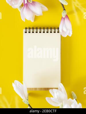 Flat lay composition with an open notebook with a white blank sheet of paper and branches of a blooming magnolia on a sunny, bright yellow background Stock Photo