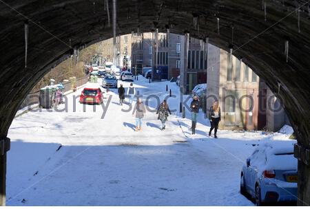 Edinburgh, Scotland, UK. 11th Feb 2021. Large icicles hanging from the underside of a bridge and People enjoying a Glorious sunny but freezing day on a snow covered Stockbridge Street. Credit: Craig Brown/Alamy Live News Stock Photo