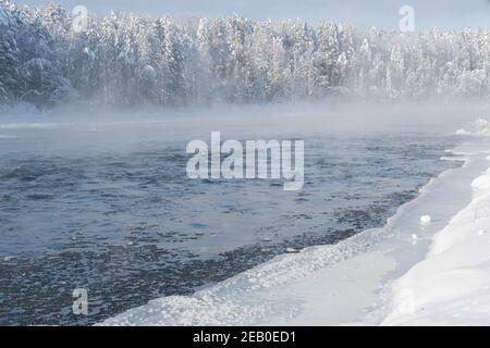 Frosty fog over winter river with snow and forest on bank. First ice on lake on cold day. Stock Photo