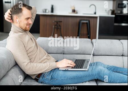 Side view a guy is using laptop lying on the comfortable couch, a young man watching movies, web surfing, play video games. A freelancer is works Stock Photo
