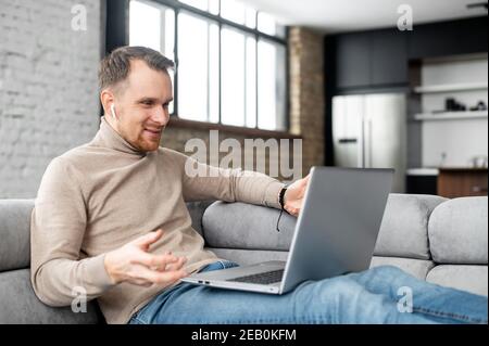 A smiling bearded hipster man is sitting on the comfy couch in the living room, relaxing and talking to his friend or family on the laptop, staying Stock Photo