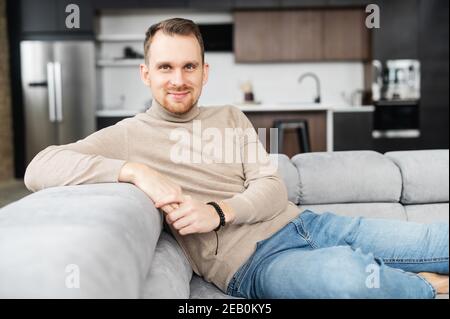Portrait of handsome young man having rest in modern living room at home, sitting casually on sofa, looks at the camera, feeling relaxed. Lifestyle Stock Photo