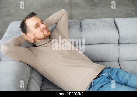 Handsome smiling guy take a nap lying on the comfortable sofa with eyes closed and hands behind head, attractive bearded man rests, daydreaming Stock Photo