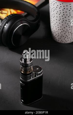 Refillable vape pod, headphones, and a plastic coffee cup on the dark surface Stock Photo