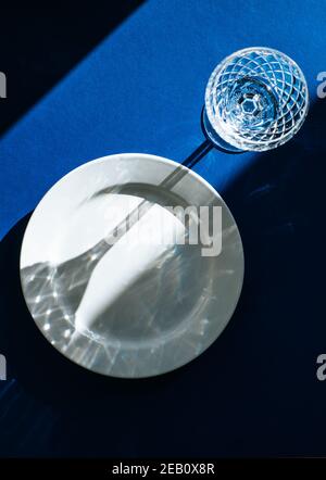 Table Setting with empty white plate and crystal glass isolated on dark blue background, dark shadows and sunlight reflecting through the wine glass Stock Photo