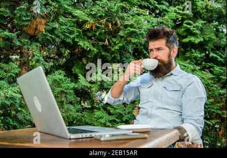 Guy drinks coffee relaxing terrace branches background. Man bearded hipster make pause for drink coffee and relax while sit with laptop. Take moment to enjoy life. Aroma cappuccino. Pleasant moment. Stock Photo