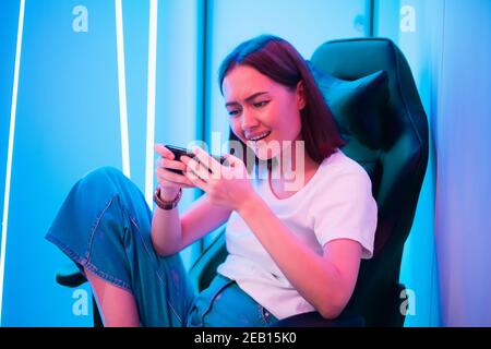 Young female professional cybersport gamer play mobile game by smartphone on eSport tournament online in colorful neon lights room. Stock Photo