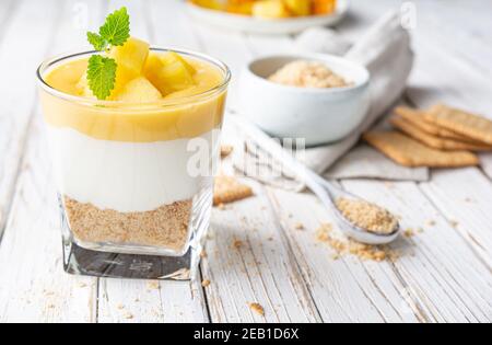 Sweet layered dessert with crushed graham crackers, cream cheese and pineapple curd, topped with fresh fruit slices in a glass jar on old wooden backg Stock Photo