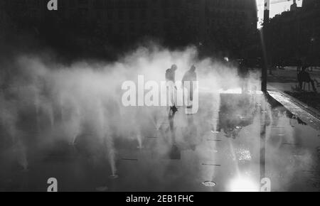 Blurry silhouettes of unrecognizable boy and girl playing in fountain water mist at autumn day. Les Halles square, Paris, France. Black and white phot Stock Photo