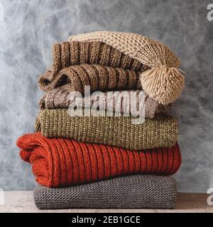 Stack of folded knitted wool women sweaters, scarf, hat with pompom on gray background. Season of warm knitwear. Fashion concept Stock Photo