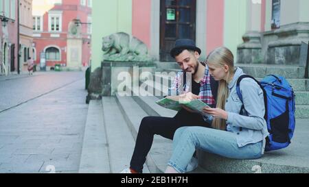 Happy tourist couple sitting on street stairs, checking city map and discussing. In the background there is beautiful old town view. Stock Photo