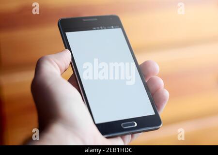 Closeup of male hand holding modern black mobile phone with blank screen for graphic designs isolated on brown blurry background. Stock Photo