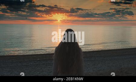 Close-up shot of woman with long hair walking to the sea and raising her hands up. Looking on sunset. Back view.