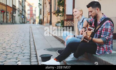 Young man and woman of street singers sitting on sidewalk, playing guitar and singing. Lifestyle concept. Stock Photo