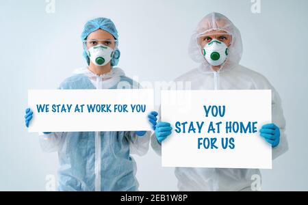 Medical workers wearing medical masks amd protective suits, holding paper with text 'WE STAY AT WORK FOR YOU, YOU STAY AT HOME FOR US. Stock Photo
