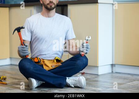 Serving you. Cropped shot of young handyman holding a hammer and an adjustable wrench in his hands while sitting on the floor indoors Stock Photo
