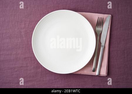 Clean empty white plate, fork and knife on bright blueberry red linen tablecloth on table, mockup Stock Photo
