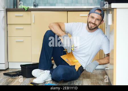 Fix it right. Full length shot of young handyman, plumber wearing tool belt sitting on the floor and smiling at camera while examining a sink before Stock Photo