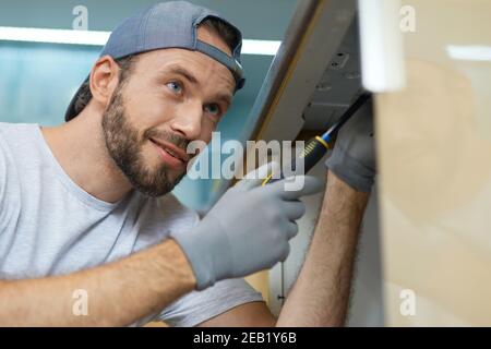 Dedicated to excellence. Close up portrait of young repairman, professional plumber looking cheerful while fixing a sink using screwdriver in the Stock Photo