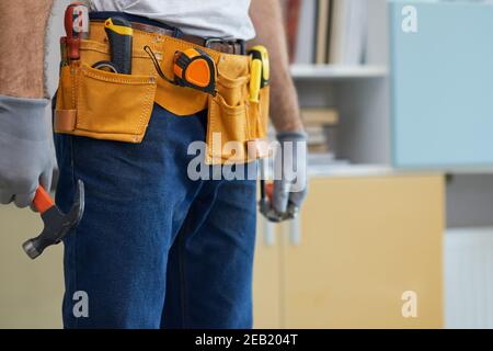 Cropped shot of professional repairman wearing a tool belt, holding a hammer and an adjustable wrench in his hands while standing indoors Stock Photo