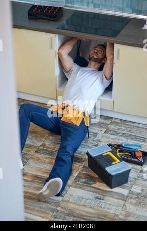 Stop leakage. Professional repairman, plumber wearing tool belt examining and fixing sink pipe in the kitchen indoors Stock Photo